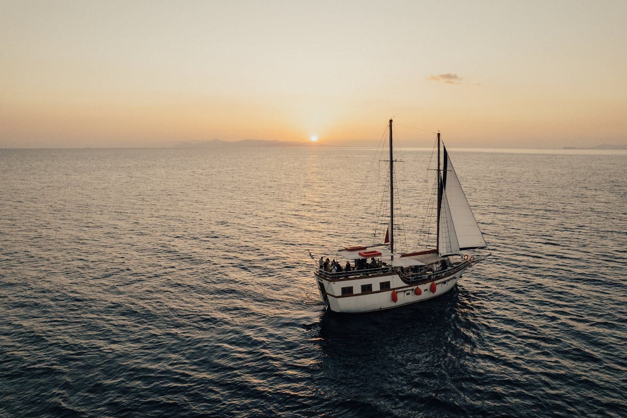 White sailboat cruising at sunset on a calm sea with distant mountains on the horizon.