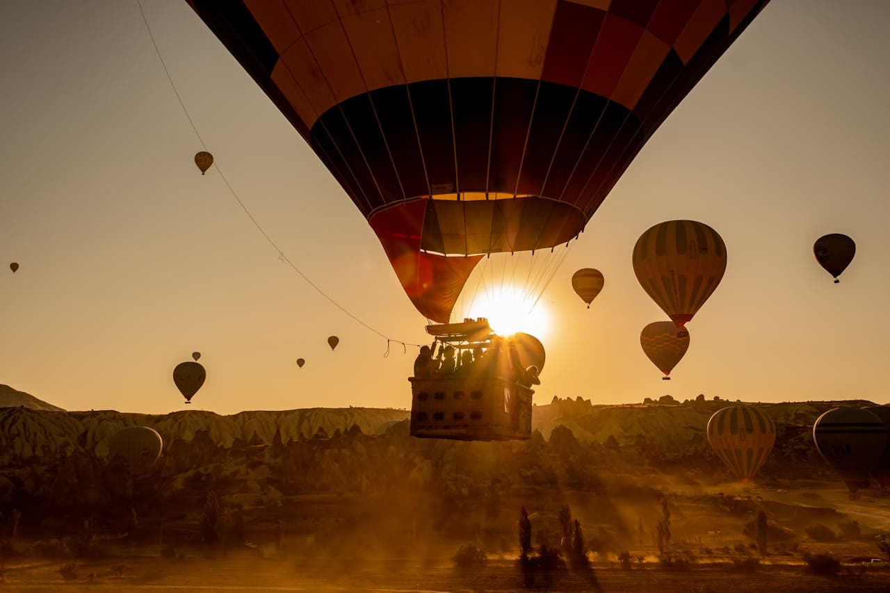 Hot air balloons float above a sunlit landscape, with one prominently silhouetted against the rising sun, creating a warm and scenic view.