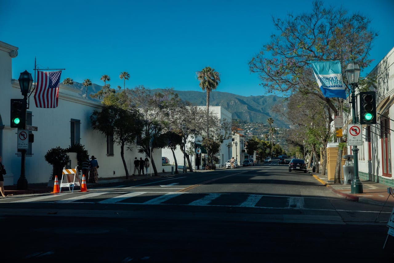 Street scene with traffic lights, American flag, trees, and mountains in the background under a clear blue sky. Crosswalk and road signs visible.