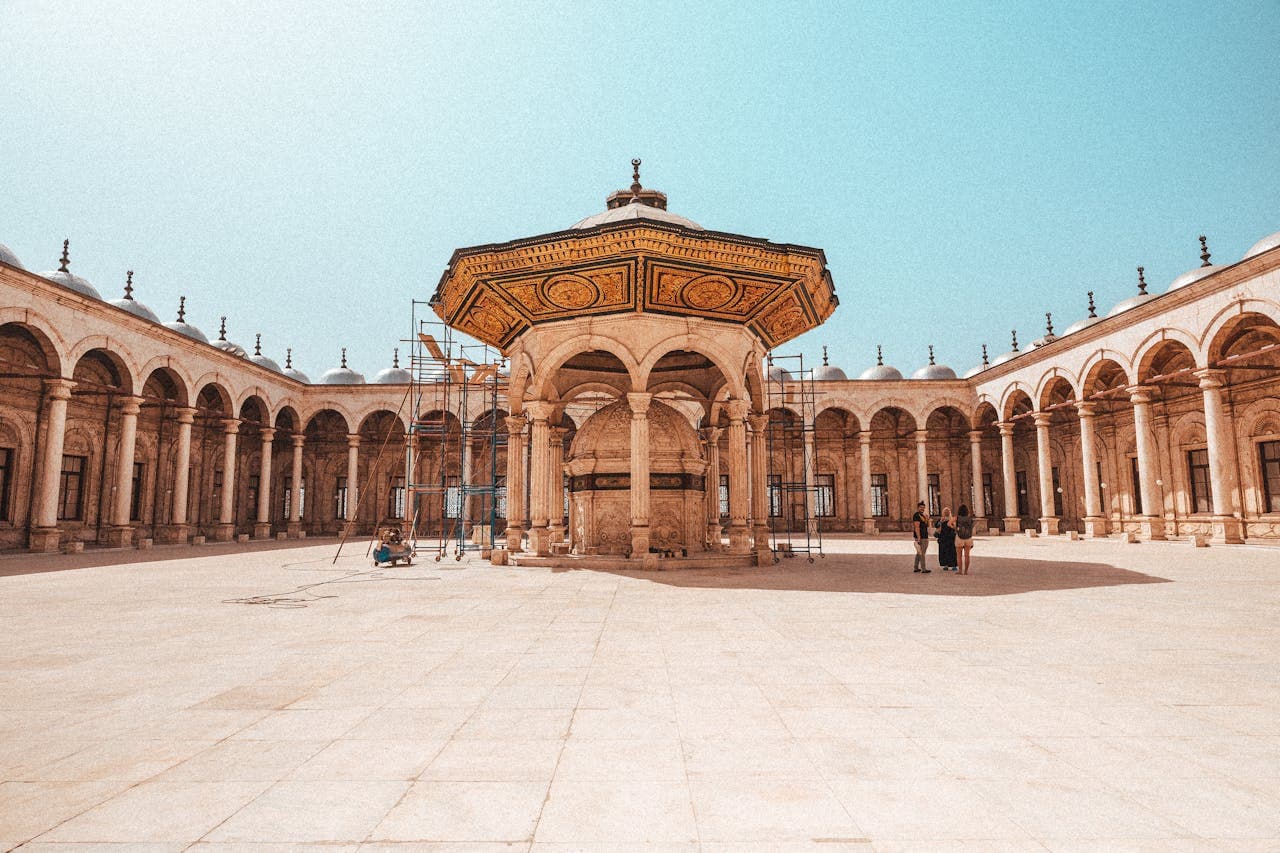Interior courtyard of the Mosque of Muhammad Ali in Cairo, Egypt, featuring the ornate central ablution fountain surrounded by arcaded walkways and marble columns under a clear blue sky.