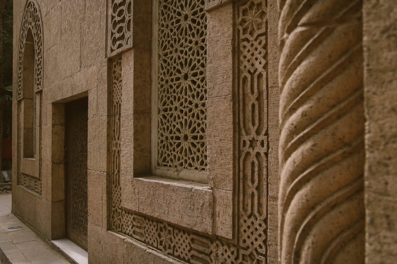 Close-up of an intricately carved stone wall featuring geometric patterns and decorative window grilles. The craftsmanship highlights traditional architectural motifs, with a doorway and window set into the textured stone surface