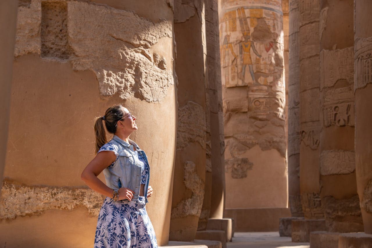 A woman stands among the massive, ancient columns of an Egyptian temple, gazing upward at the hieroglyphs and carvings on the stone pillars, with sunlight illuminating the scene.