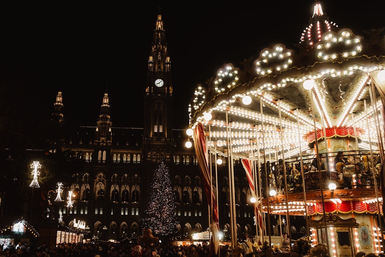 Festive nighttime scene with a brightly lit carousel, a decorated Christmas tree, and an illuminated historic building in the background.