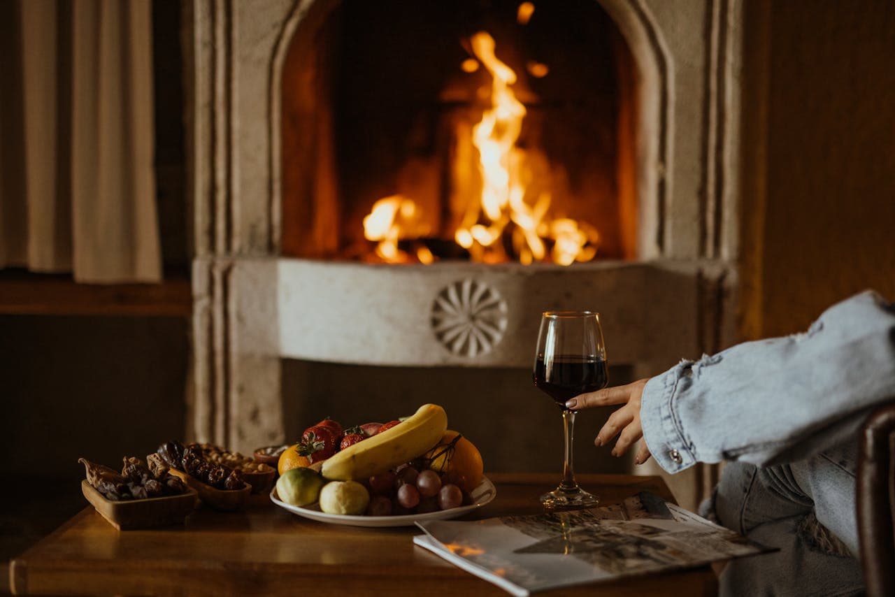 Cozy fireplace setting with a plate of fruits, nuts, a glass of red wine, and a person's hand reaching for the glass on a wooden table.