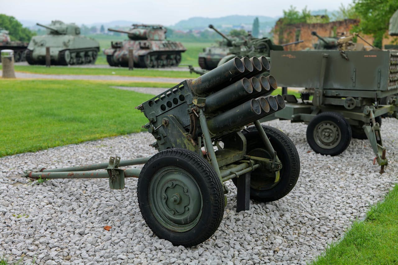 Historic military artillery and tanks displayed outdoors at a military museum, featuring green armored vehicles and rocket launchers on a gravel path with grassy surroundings.