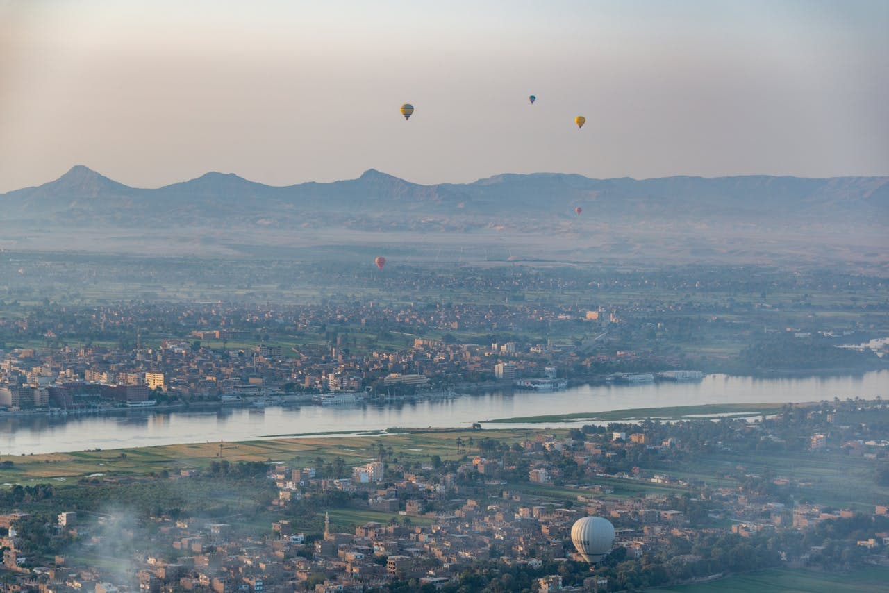A panoramic aerial view of a city along the Nile River, with hot air balloons floating in the sky and green fields, buildings, and distant mountains visible in the background.