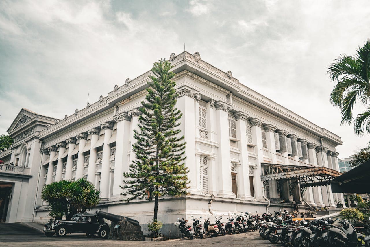 Historic white neoclassical building with columns and ornate details, surrounded by motorcycles and tropical trees on a cloudy day.