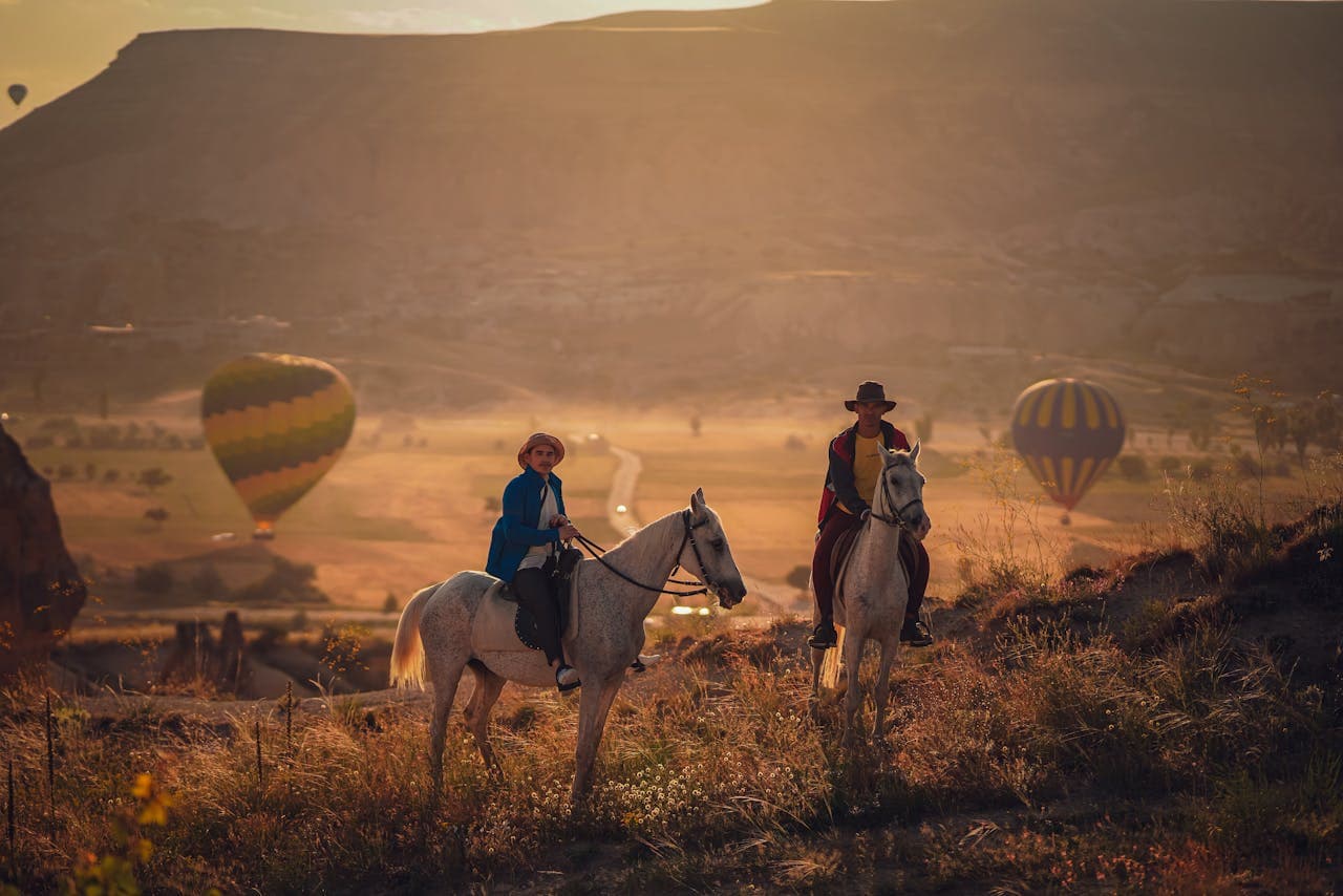 Two people riding horses in a scenic landscape with hot air balloons in the background during a warm, golden sunset.