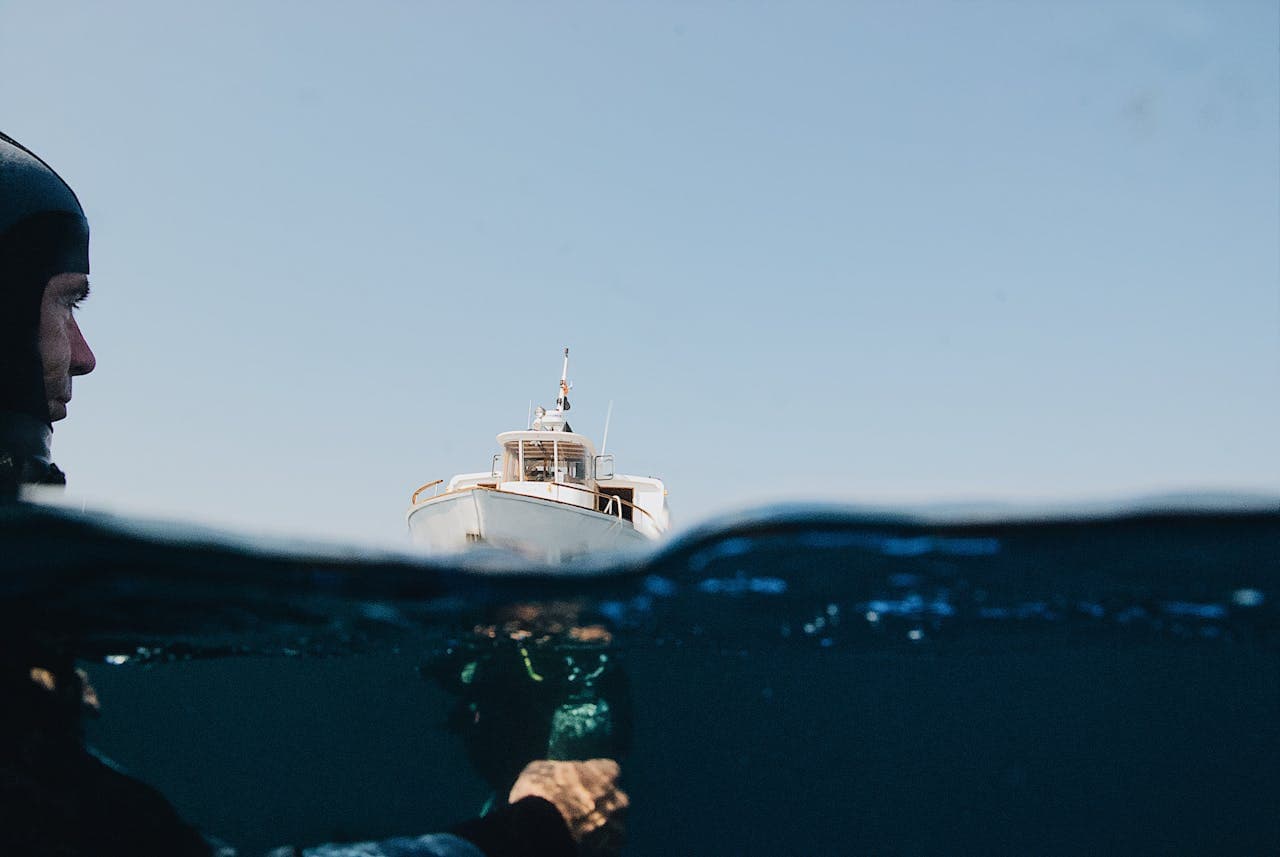 Split-level view of a diver in a wetsuit below water and a boat floating above the ocean surface under a clear blue sky.