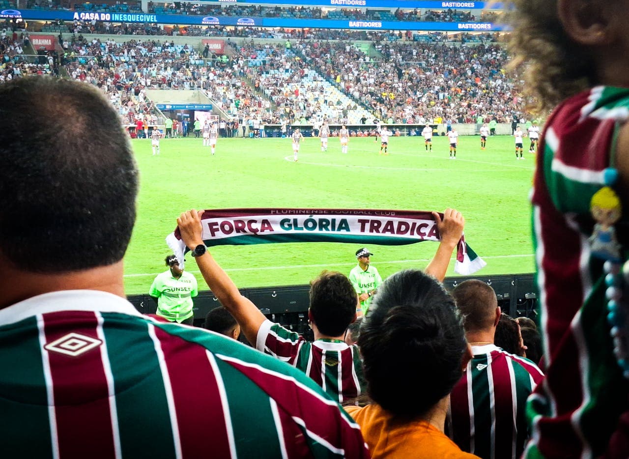 Fluminense football fans in striped jerseys cheering at Maracanã Stadium, holding a scarf that reads "Força Glória Tradição" during a live soccer match.
