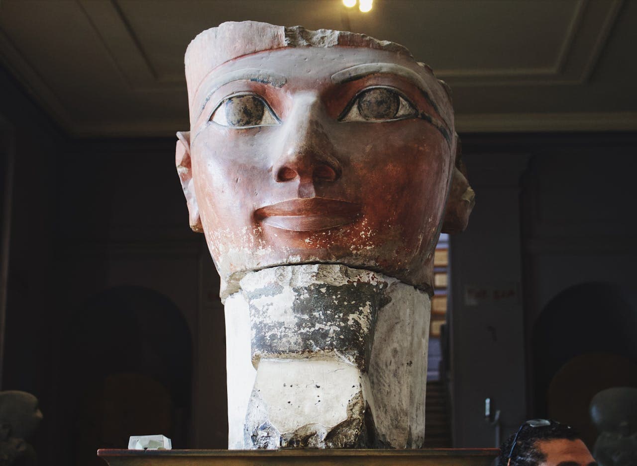 A large, weathered head of an ancient Egyptian statue displayed indoors, with a reddish-brown face, detailed eyes, and visible signs of aging and damage around the base.