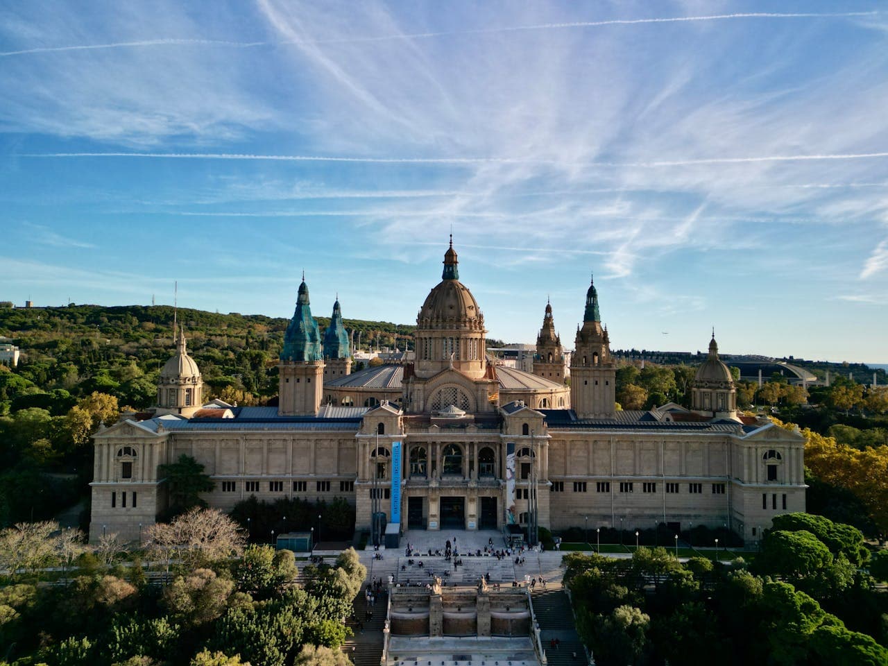 Aerial view of the National Art Museum of Catalonia (MNAC) in Barcelona, Spain, with grand architecture, domes, and lush green surroundings under a blue sky.