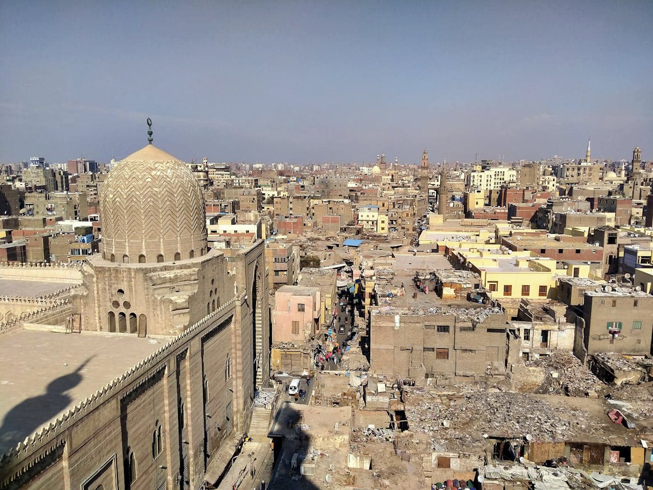 A panoramic cityscape showing a dense, historic urban area with flat-roofed buildings and narrow streets. In the foreground is a large mosque with a patterned dome and ornate façade. The background reveals a sprawling city with a mix of old and new buildings under a clear sky.