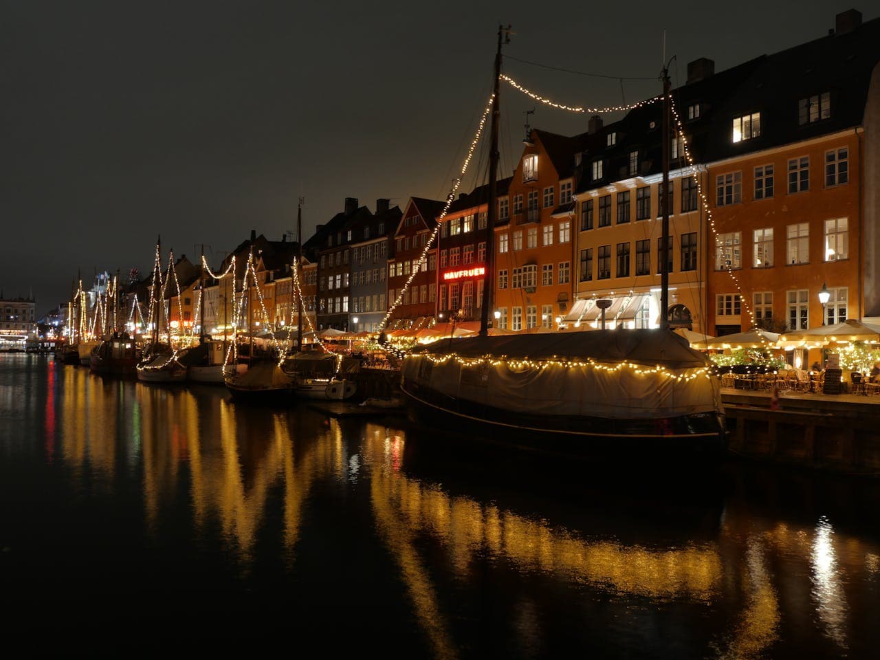 Night view of a canal lined with illuminated boats and buildings, reflecting warm lights on the water, creating a festive atmosphere.