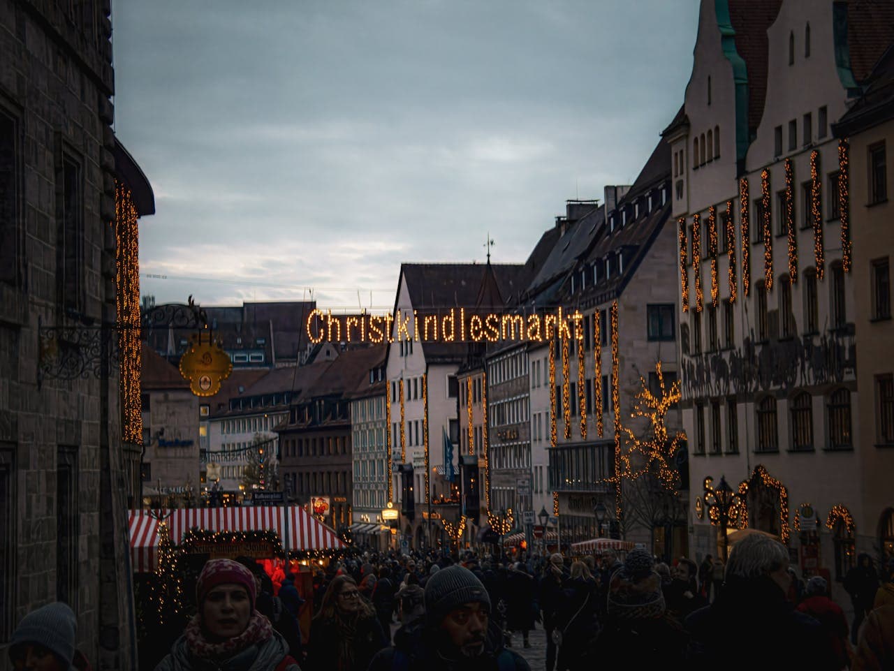 Crowded Christmas market street with festive lights and decorations, featuring a "Christkindlesmarkt" sign overhead against a cloudy sky.