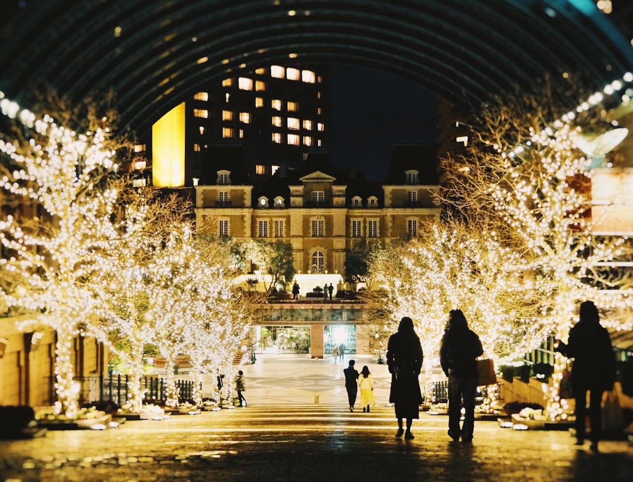 People walk under an illuminated archway lined with trees adorned with fairy lights, leading to a grand building at night.