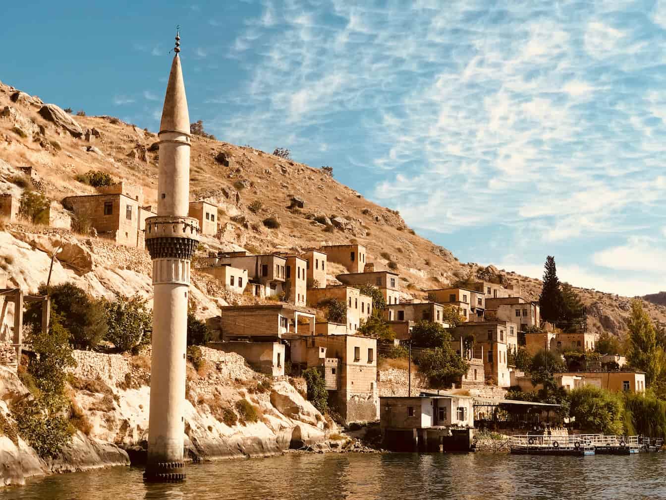 A tall minaret rises beside a lake in front of a hillside village with stone houses under a clear, partly cloudy sky.