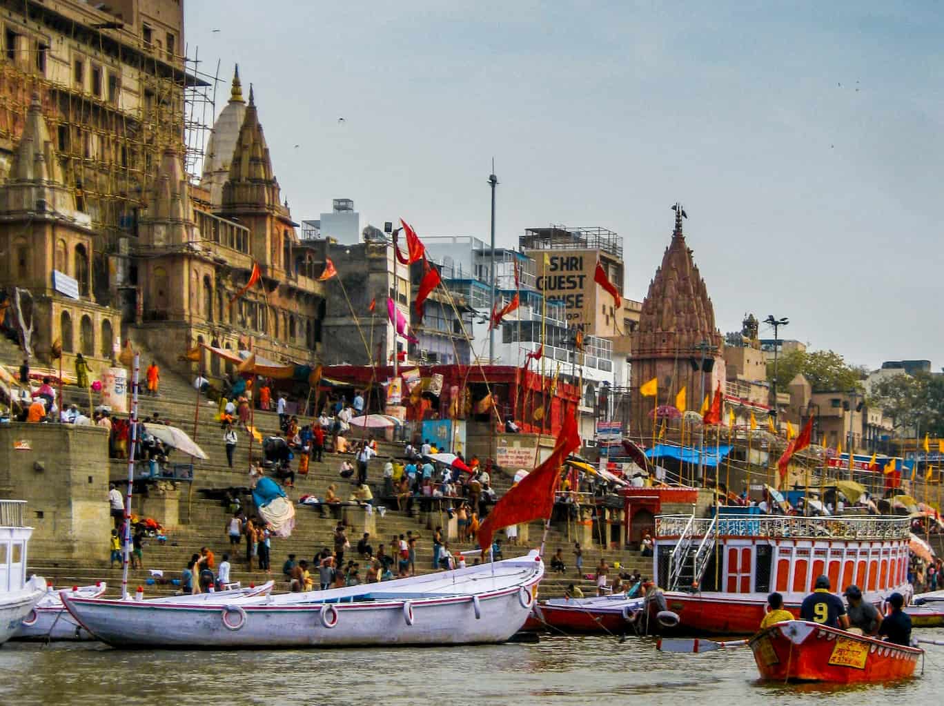 View of vibrant ghats in Varanasi, with boats on the river, bustling crowds, historic buildings, and colorful flags adorning the steps.