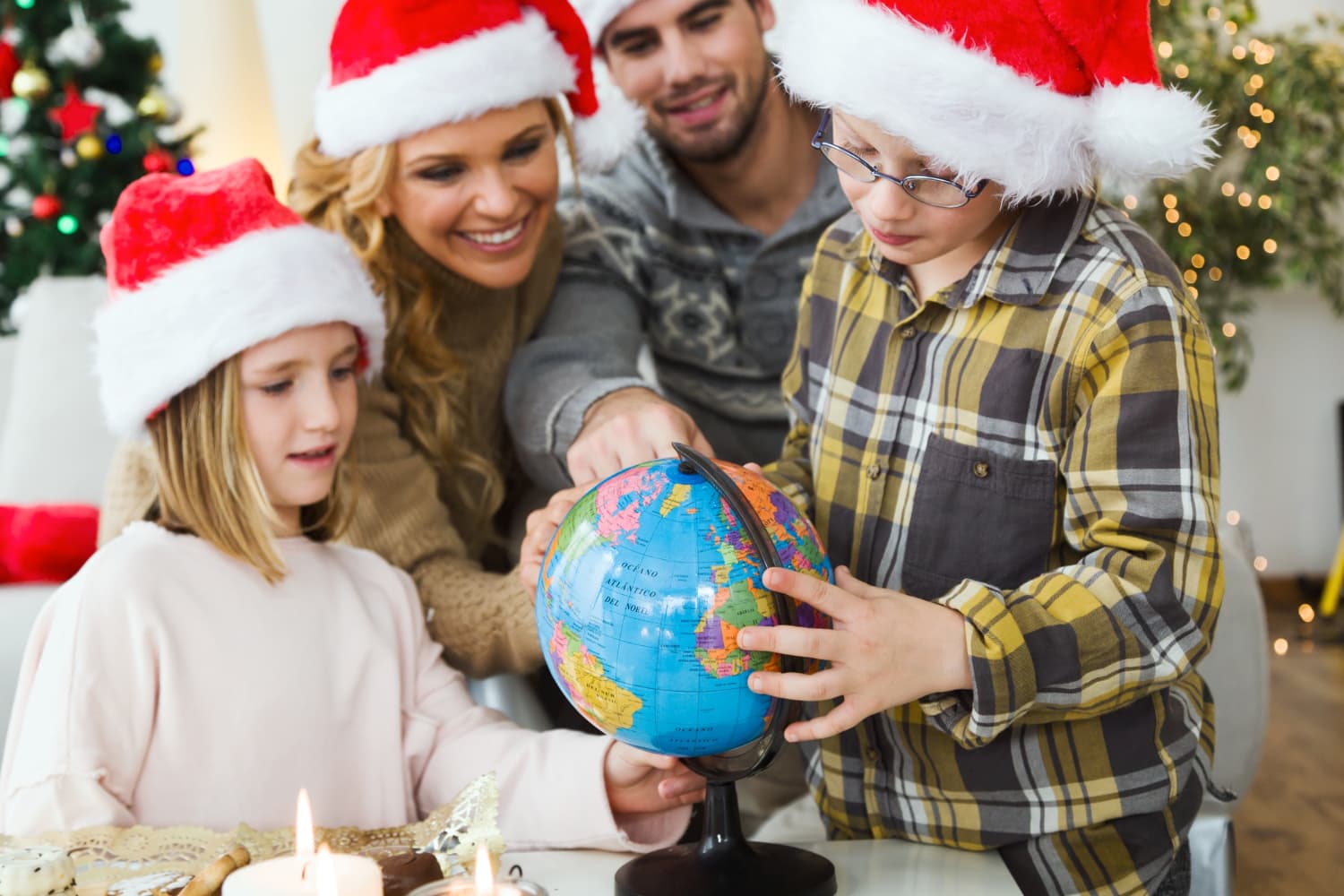 Two children wearing Santa hats examine a globe, assisted by two adults, in a festive room with a decorated Christmas tree.