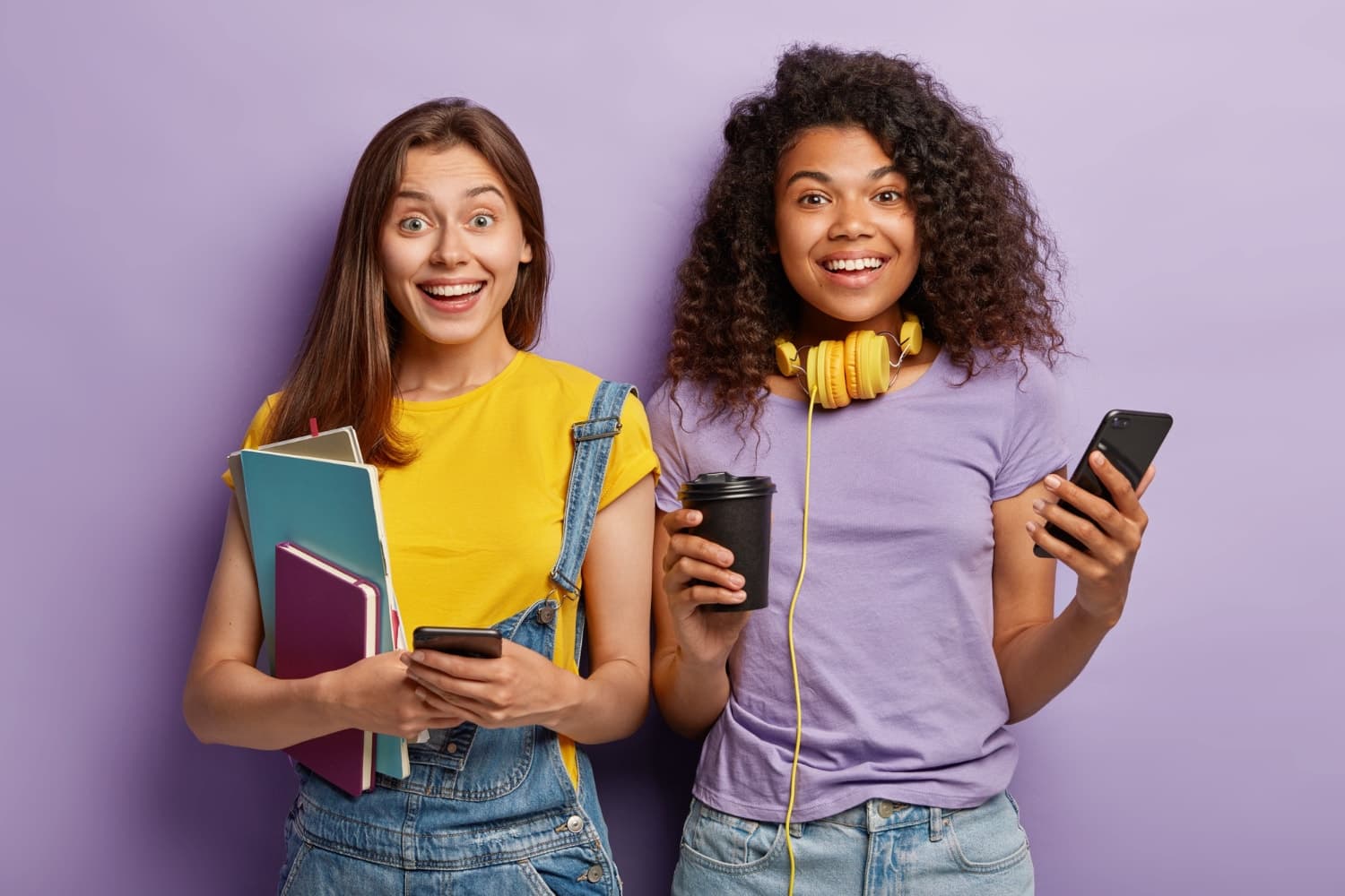 Two smiling students holding books, coffee and phones against a purple backdrop