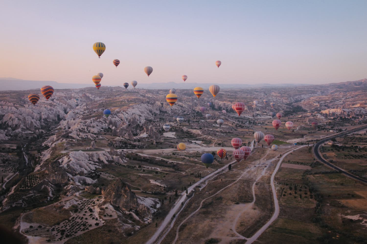 Hot air balloons in various colors float over a rocky landscape at sunrise, with winding roads and sparse vegetation below.