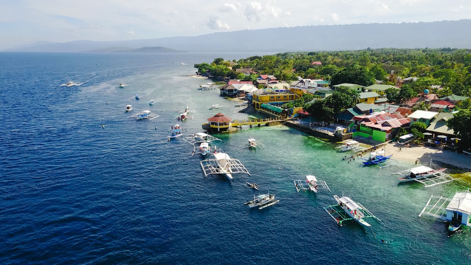 Aerial view of a coastal town with boats in clear blue water, colorful buildings along the shore, and lush green hills in the background.