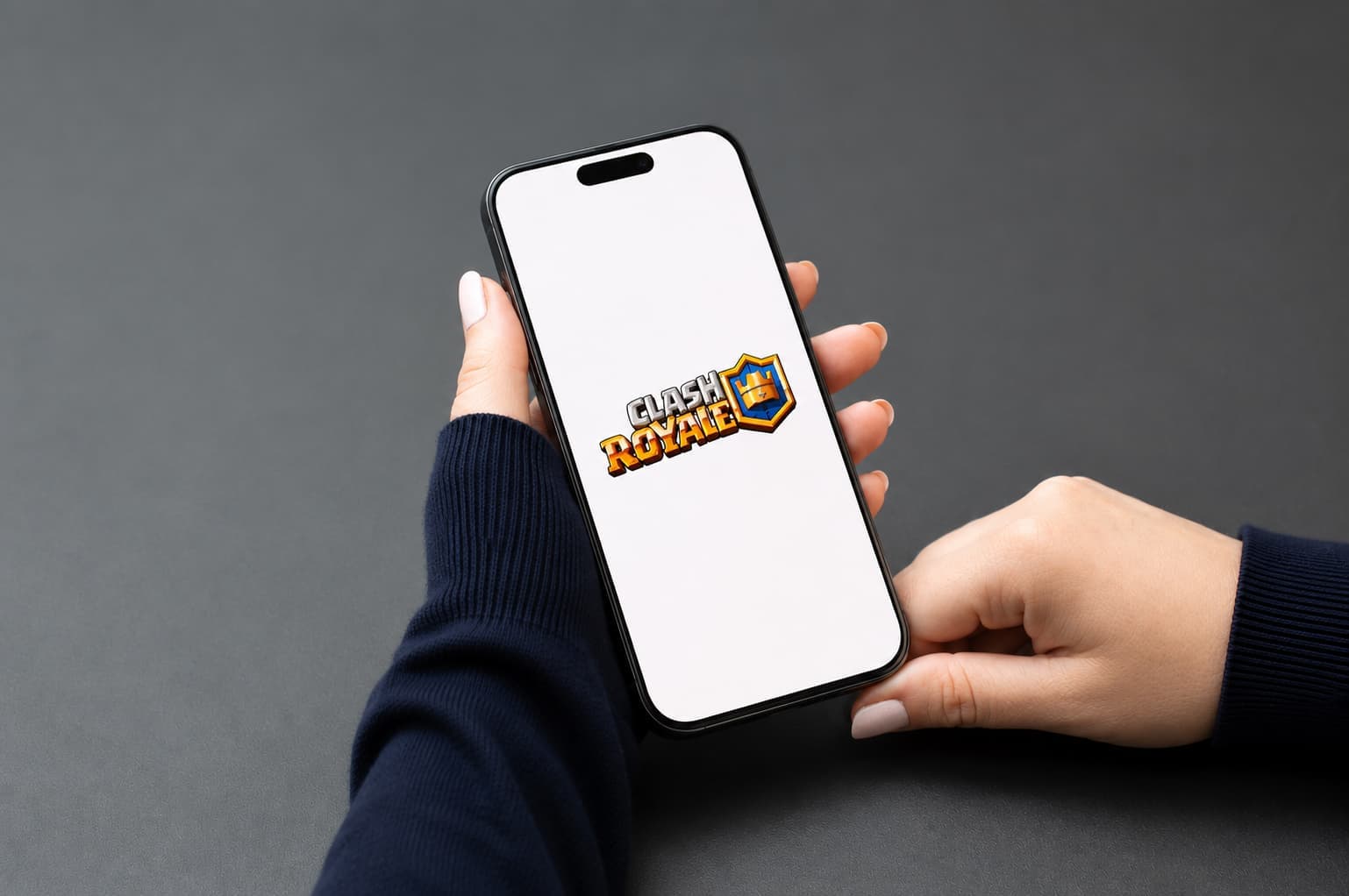Person holding a smartphone displaying the Clash Royale logo on a white screen, against a dark gray background.