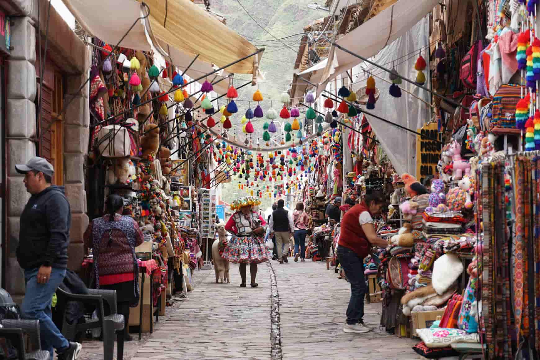 A bustling market street lined with colorful textiles, hanging ornaments, and people shopping. A woman in traditional attire stands with a llama.