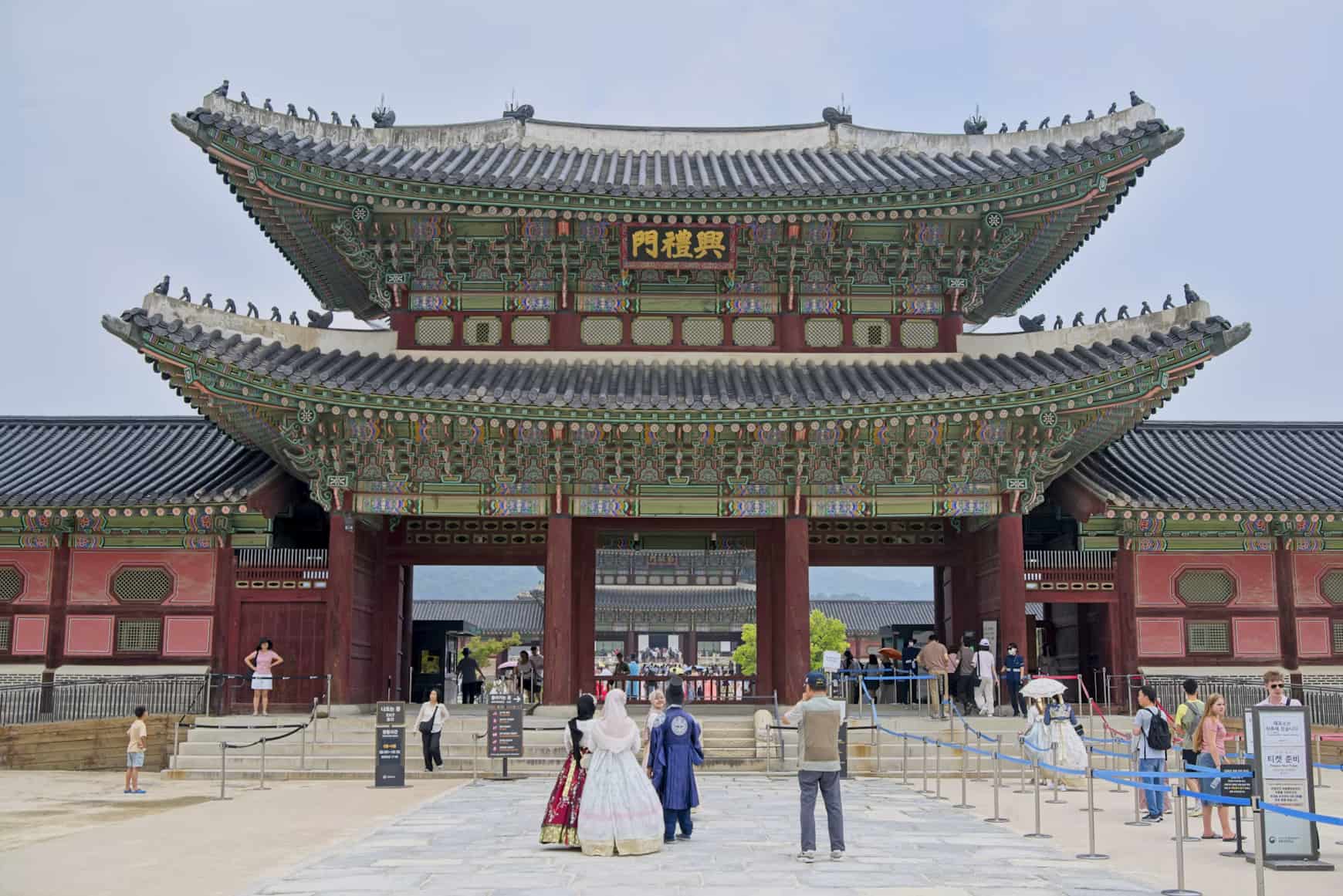 A traditional Korean palace gate with intricate designs, surrounded by visitors, some in traditional attire, under a clear sky.