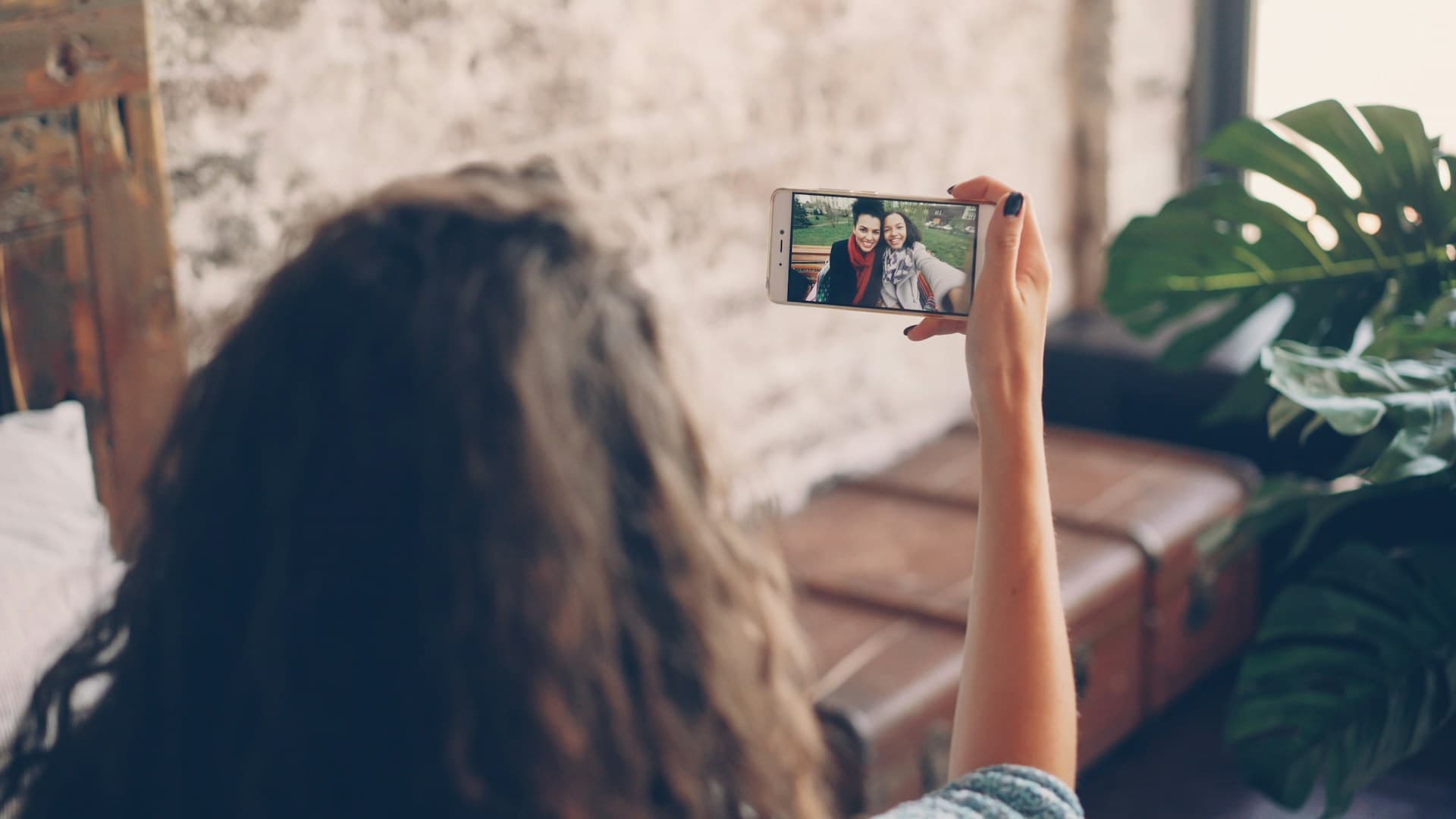 A person with curly hair is on a FaceTime call with someone else, using a smartphone. They are seated on a couch with a rustic wall and green plant in the background.