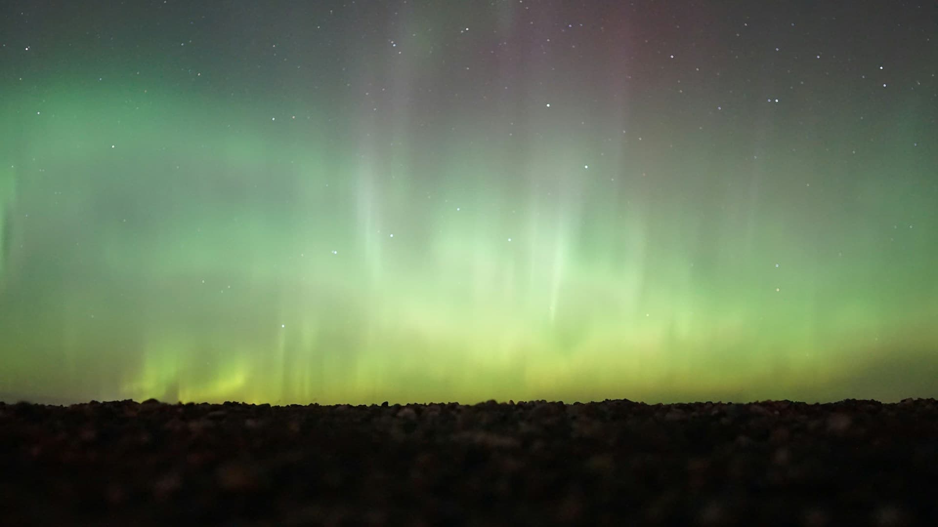 Bright green aurora borealis glowing above a dark, rocky landscape under a starry night sky.