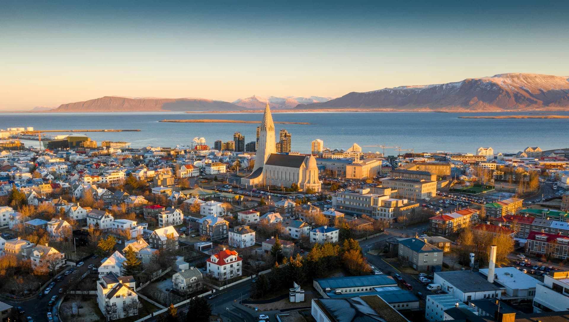 Aerial view of Reykjavik at sunset, featuring Hallgrímskirkja church, colorful buildings, and distant mountains by the sea.