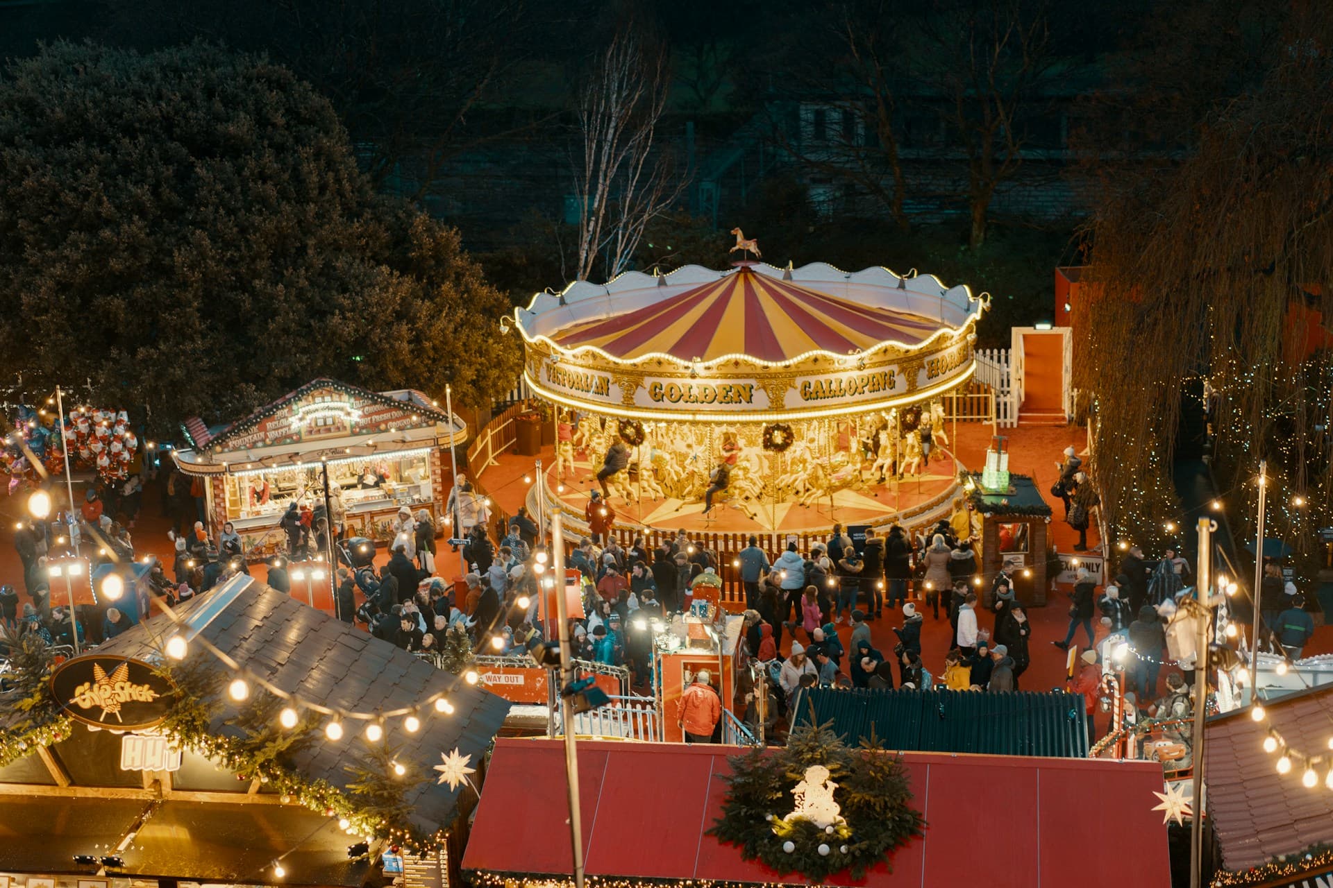 Aerial view of a festive, illuminated carousel at a bustling night market, surrounded by people and decorated stalls.