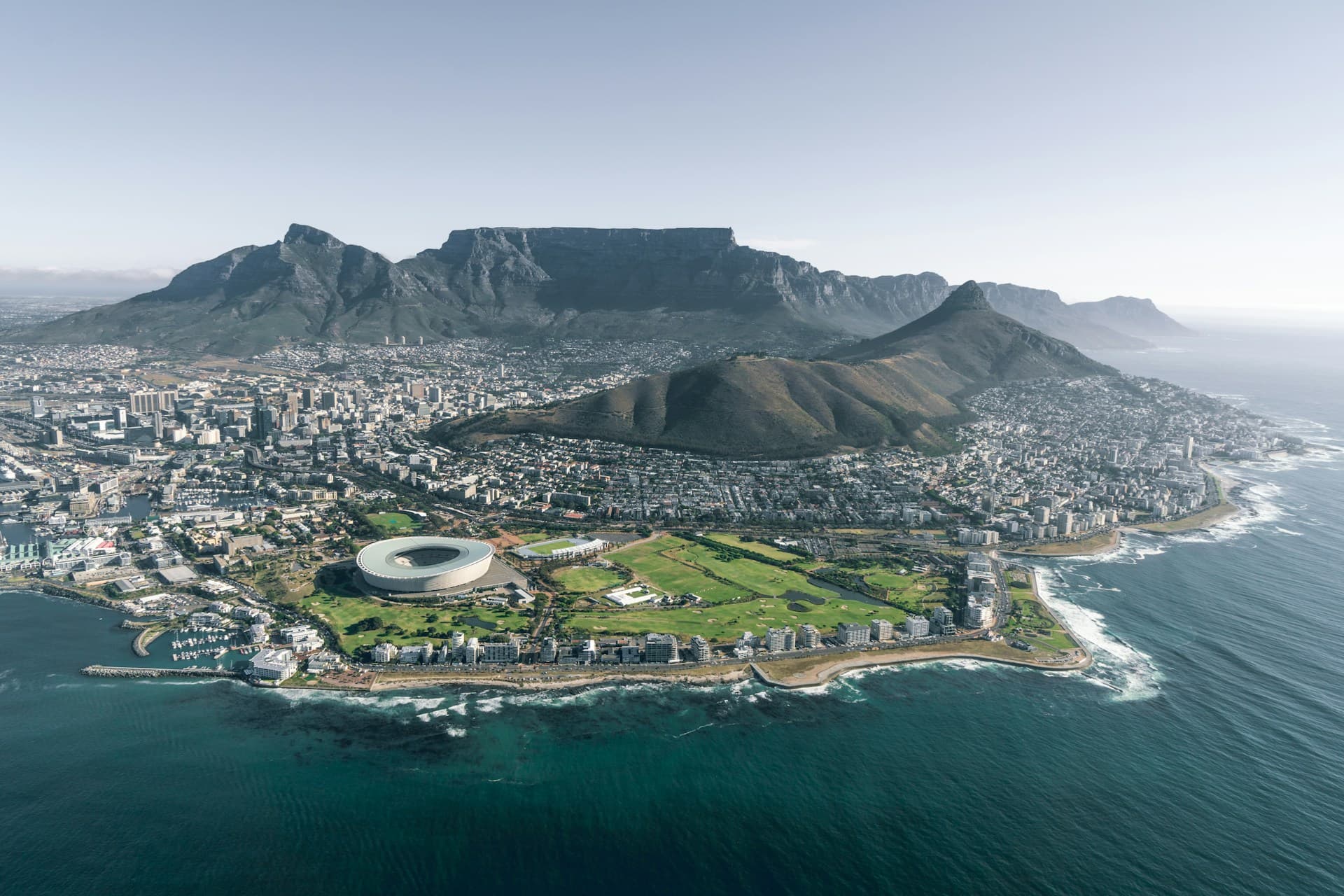 Aerial view of Cape Town, featuring Table Mountain, Lion's Head, the stadium, and coastline with clear skies and ocean waves.