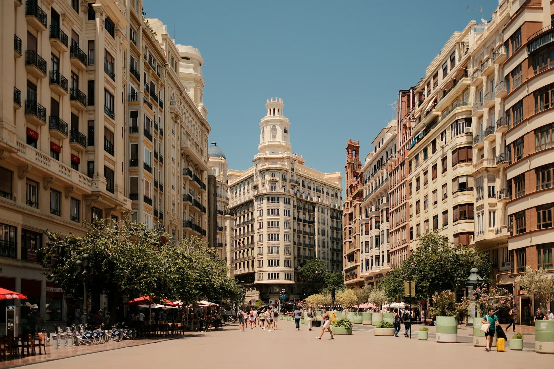Wide street view in Valencia, Spain, flanked by ornate buildings, people walking, and outdoor café umbrellas under a clear blue sky.