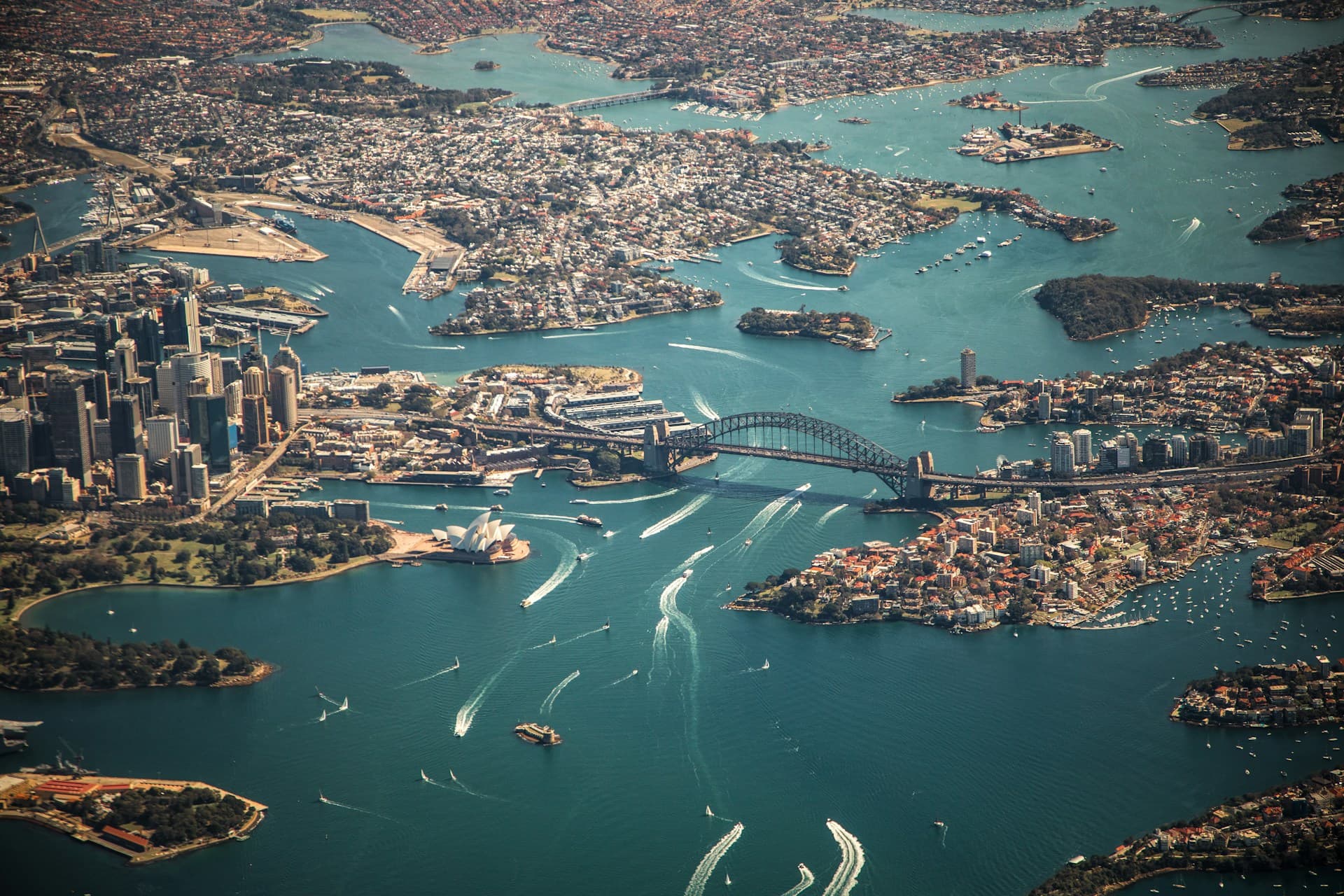 Aerial view of Sydney, featuring the Sydney Opera House, Harbour Bridge, and surrounding waterways with boats and cityscape.