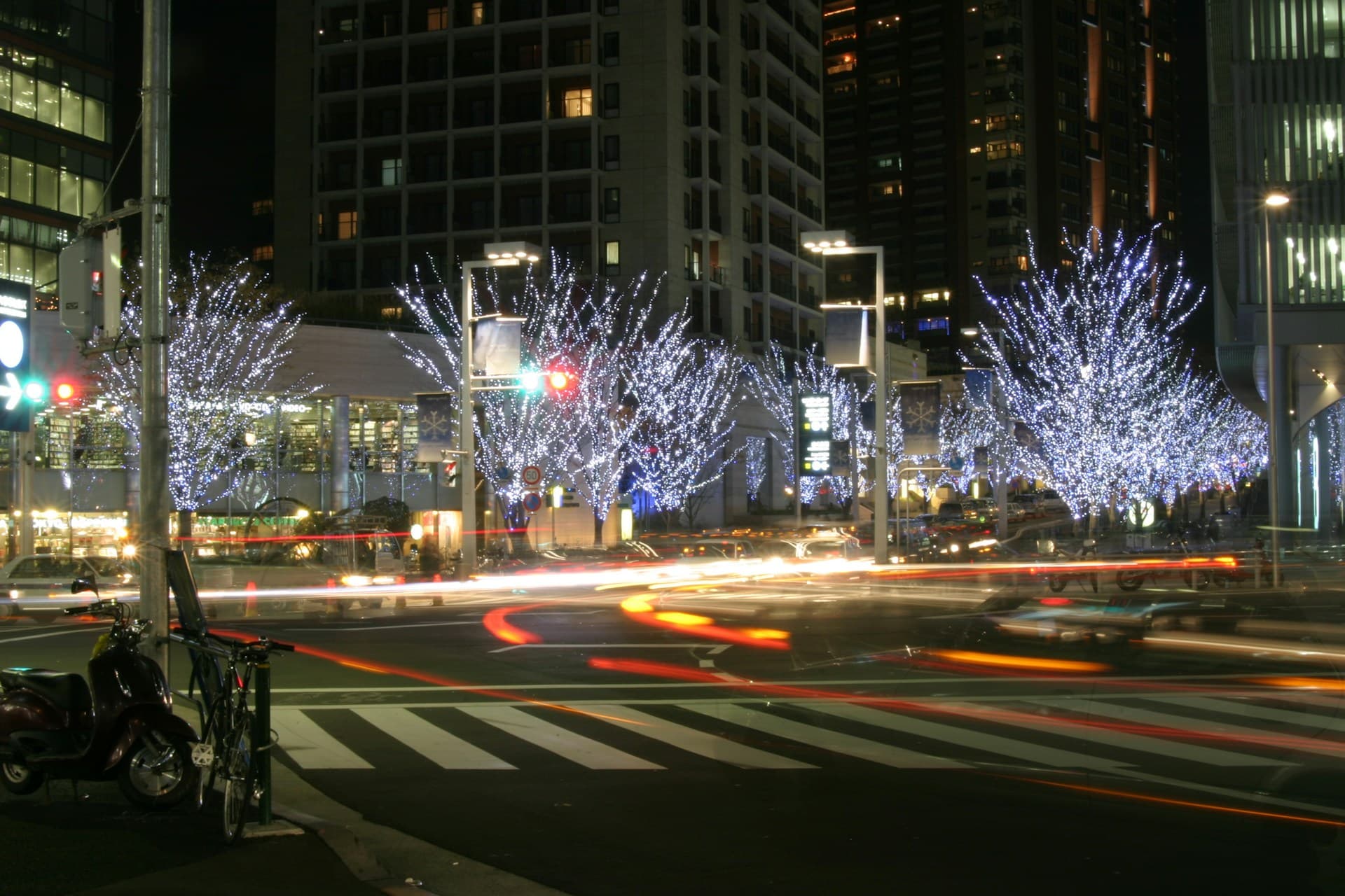 Night city intersection with trees wrapped in white lights, streaking car light trails, crosswalk, parked scooter and tall office buildings.