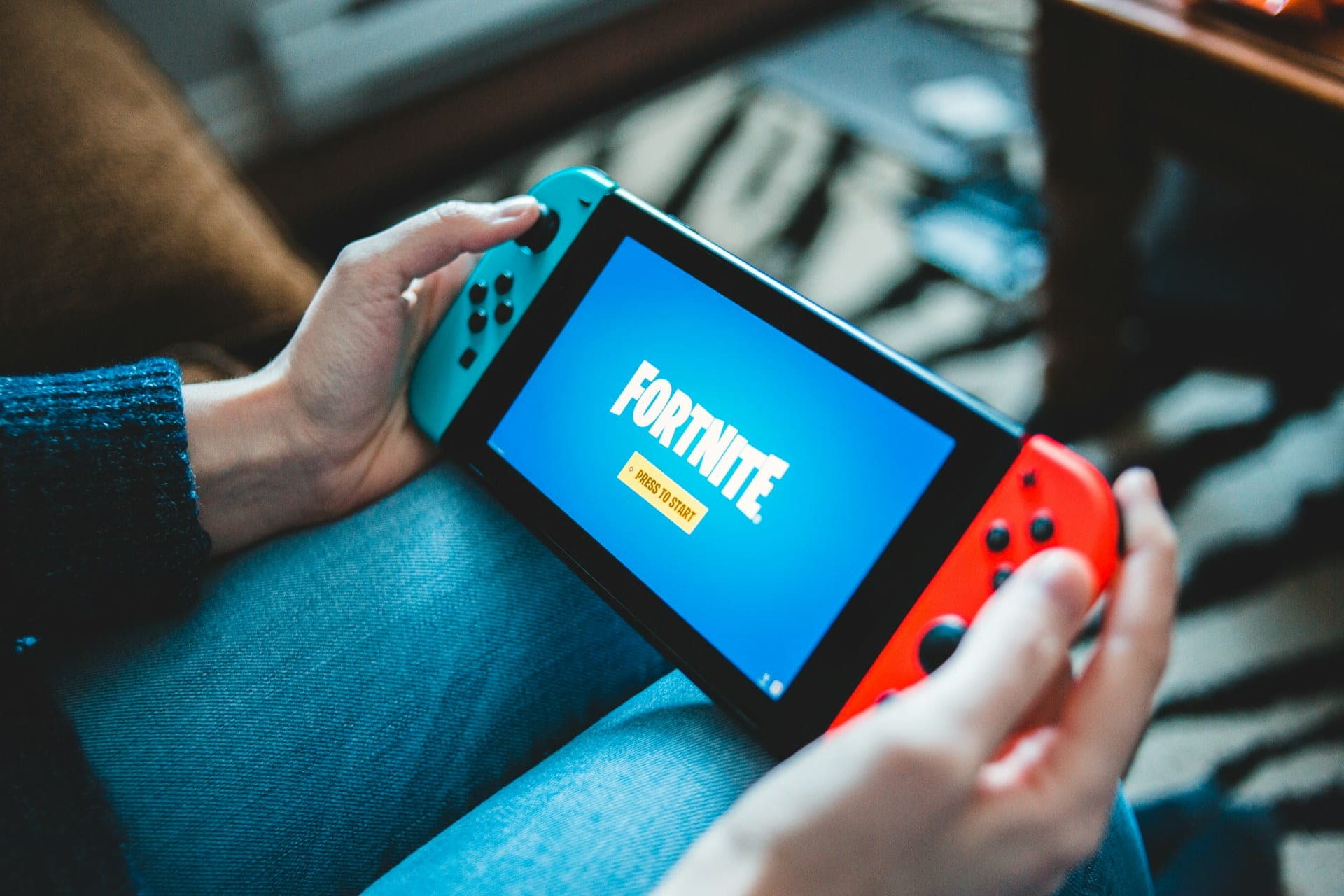 Person holding Nintendo Switch with Fortnite loading screen, seated on a couch with a patterned rug in the background.