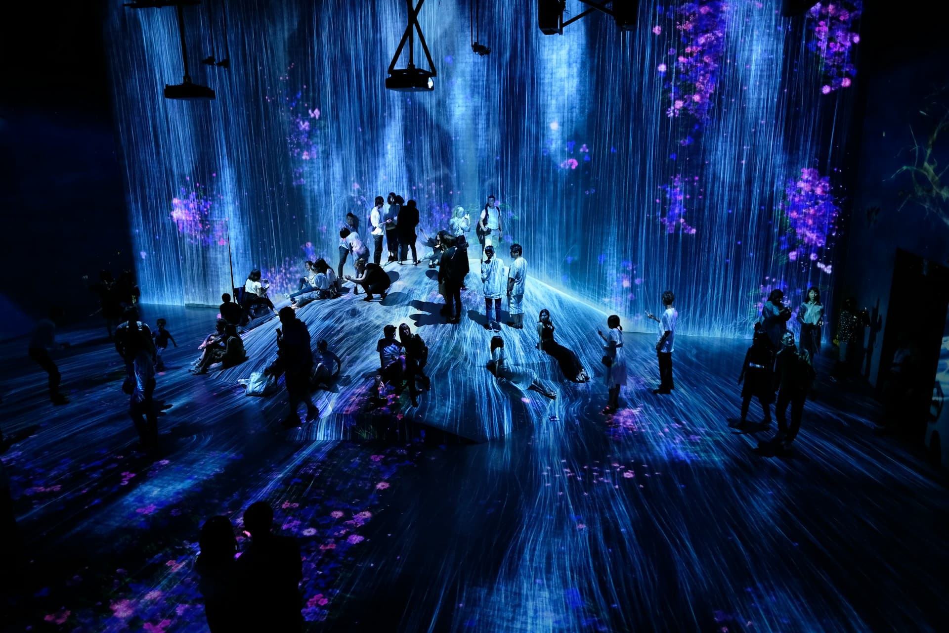 People amid cascading blue light and purple floral projections in an immersive digital art installation.