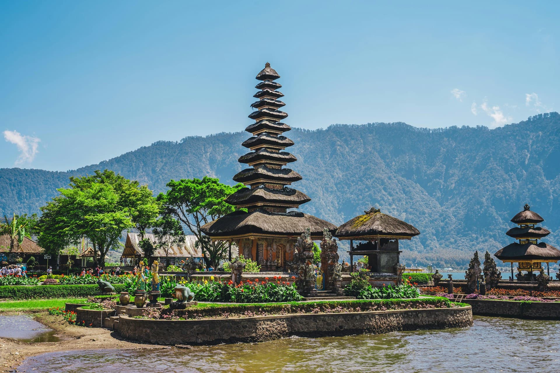 A traditional multi-tiered temple stands by a lake, surrounded by lush greenery and mountains under a clear blue sky.
