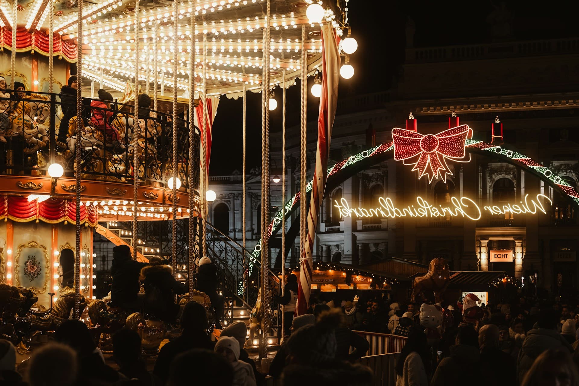 Festive Christmas market scene with a brightly lit carousel and a glowing "Merry Christmas" sign, surrounded by a crowd of people.