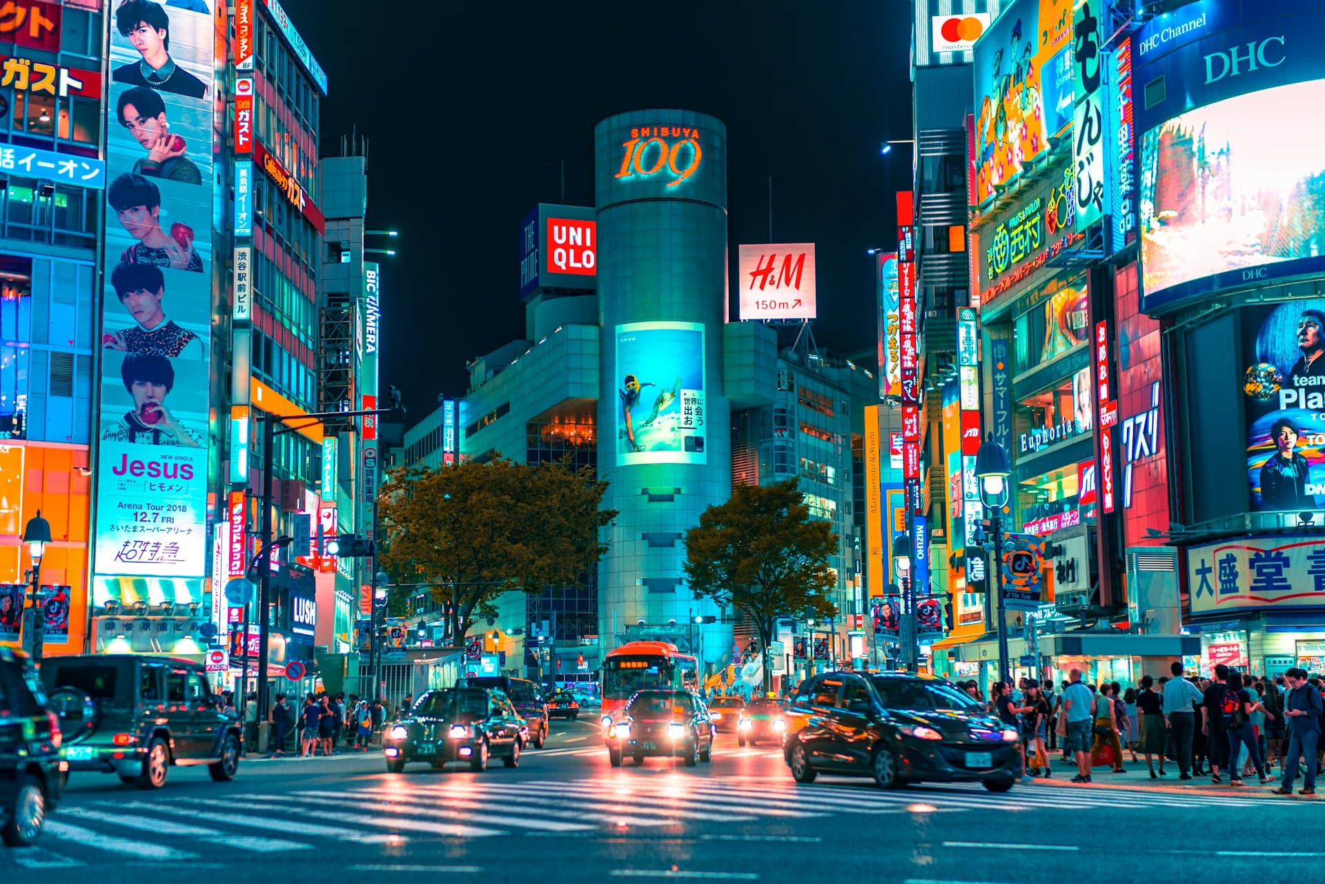 Nighttime Shibuya crossing in Tokyo, neon-lit buildings and billboards, cars waiting and crowds of pedestrians at a busy intersection.