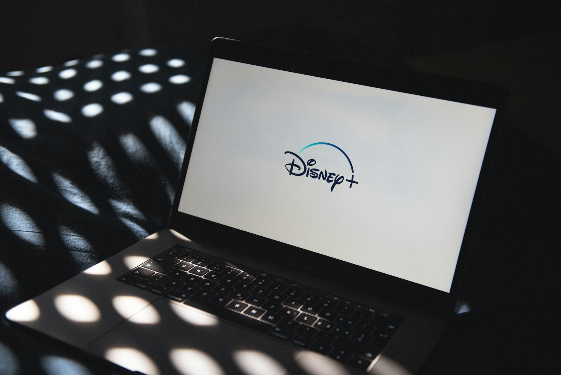 Laptop displaying the Disney+ logo on screen, with dappled sunlight creating patterns on the keyboard and surrounding surface.