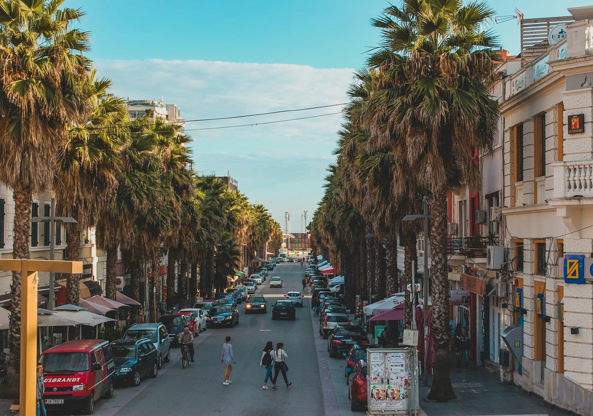 A street lined with tall palm trees, flanked by parked cars and buildings, with people crossing in the middle, under a blue sky.