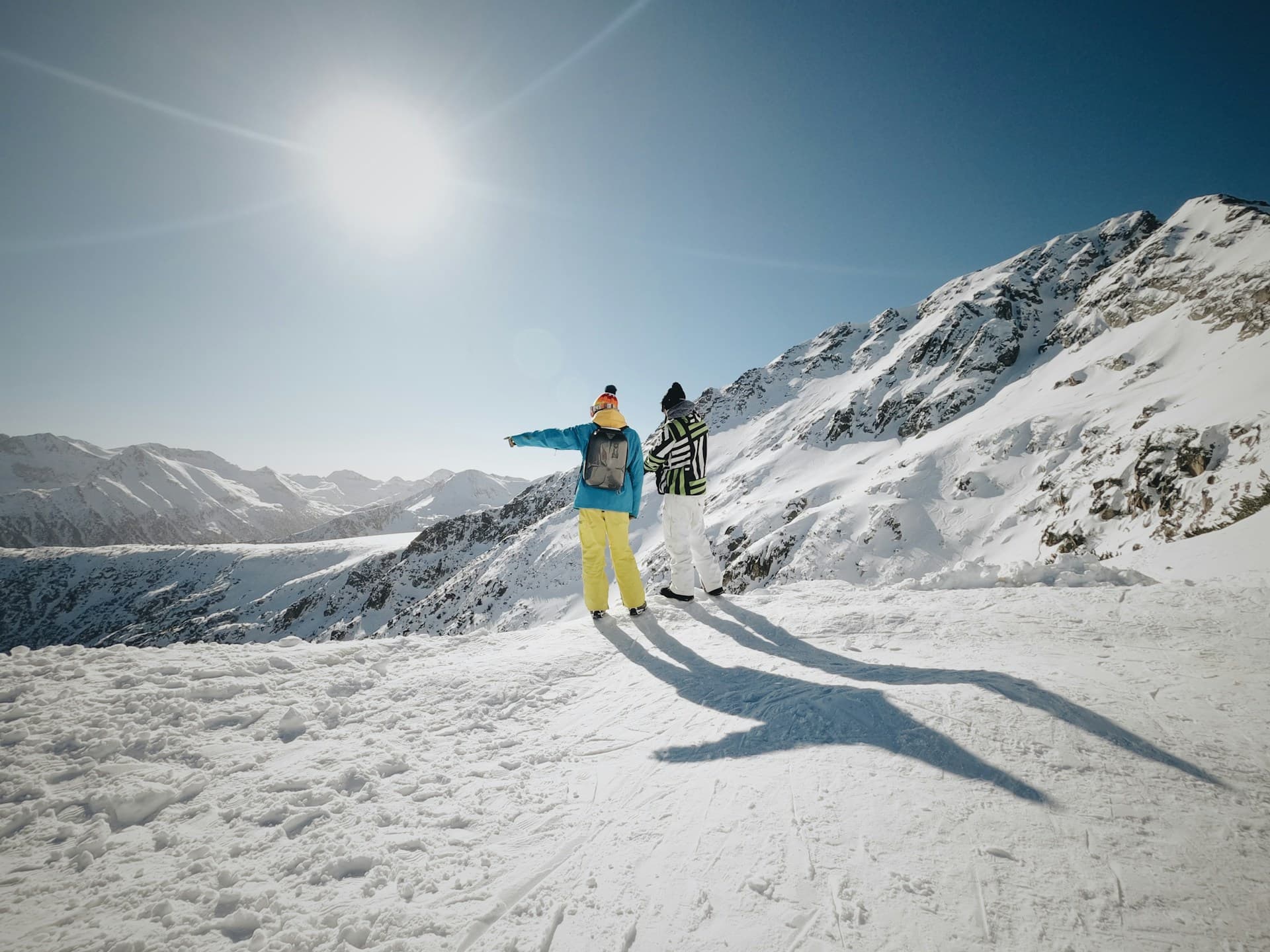 Two people in colorful winter gear stand on a snowy mountain, pointing towards distant peaks under a clear blue sky and bright sun.