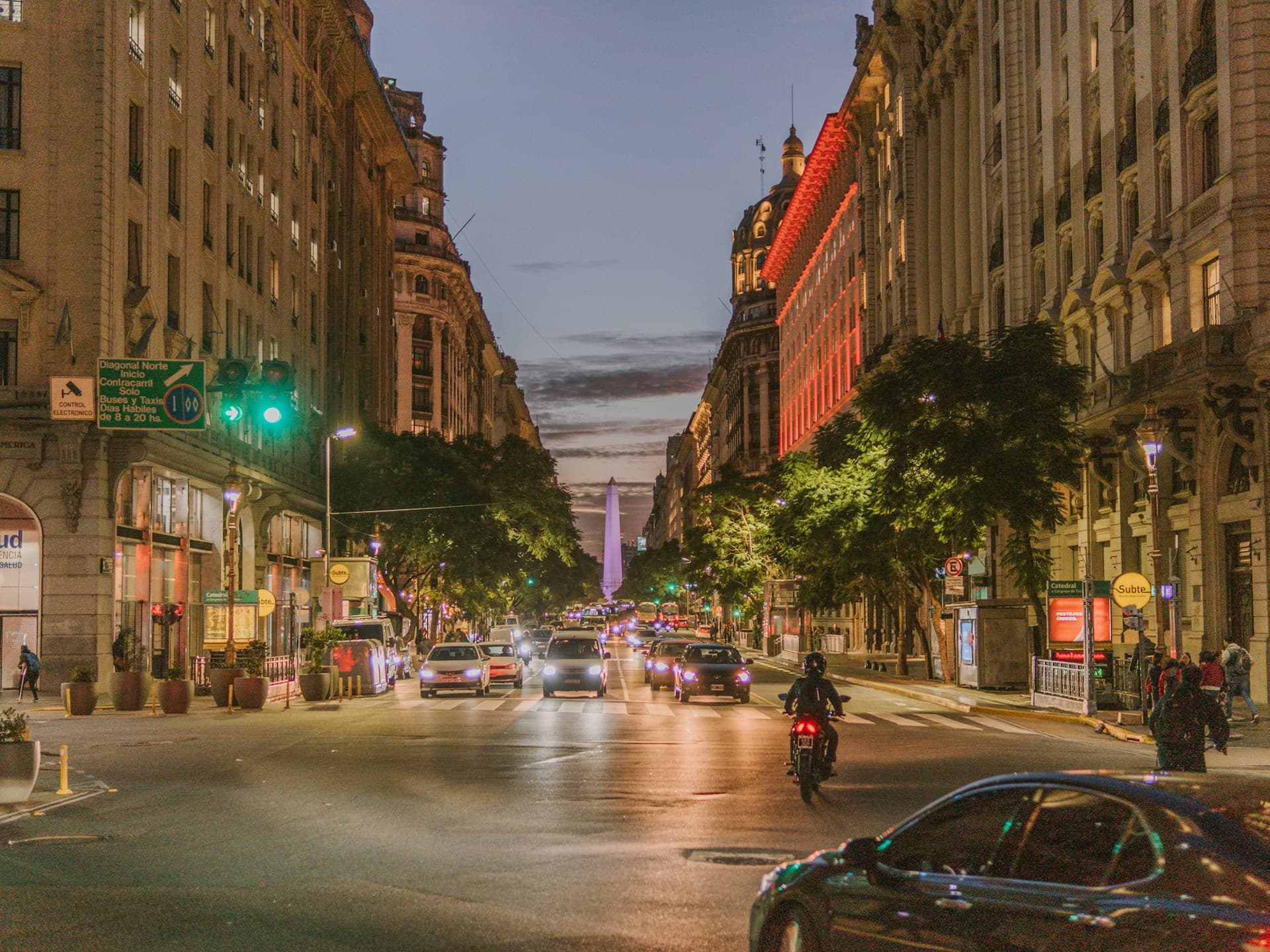 Bustling city street at dusk with cars, a motorbike, and lit buildings. A distant obelisk is visible under a twilight sky.