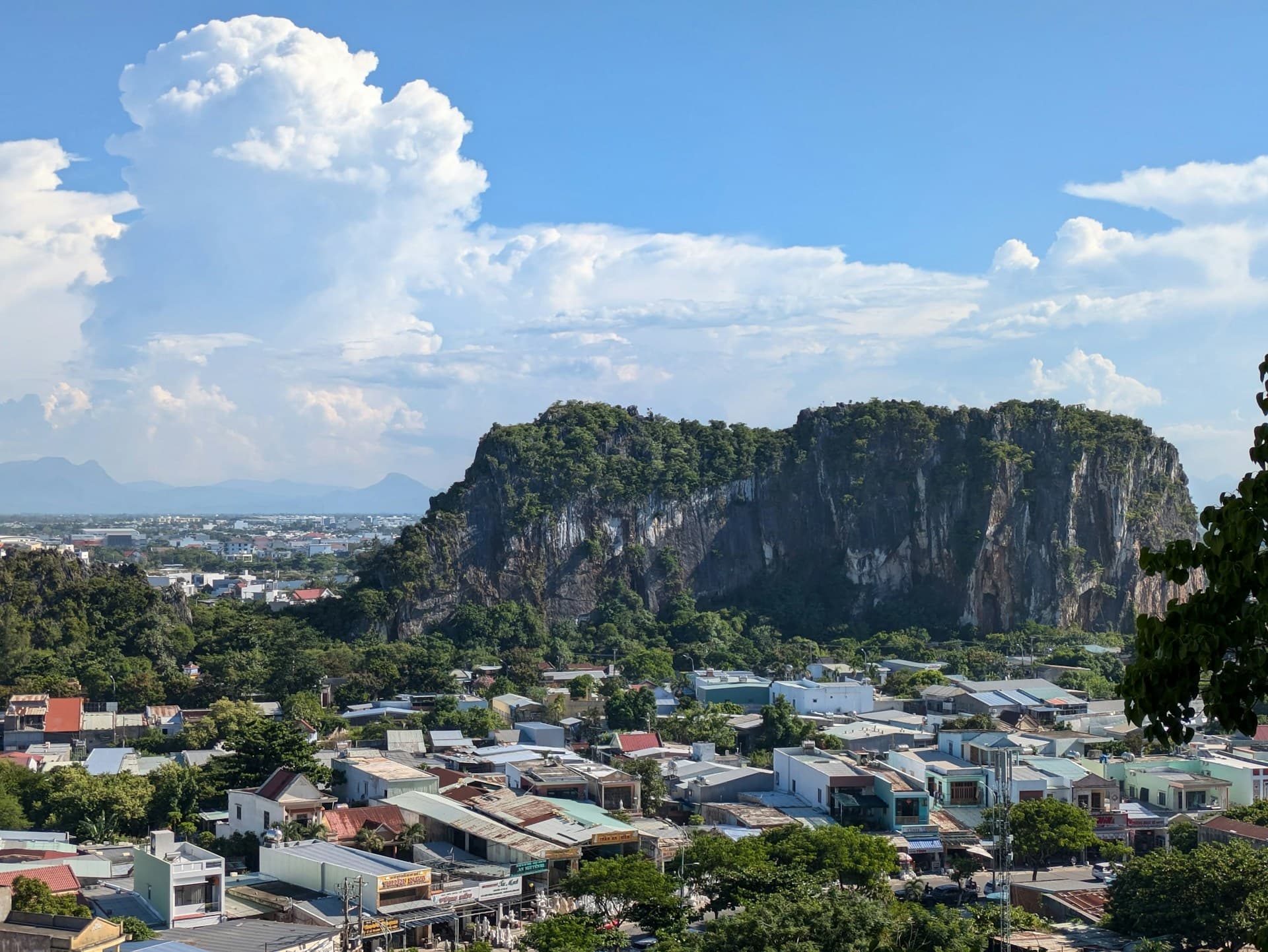 View of a cityscape with scattered buildings, lush greenery, and a large rocky hill under a blue sky with clouds.