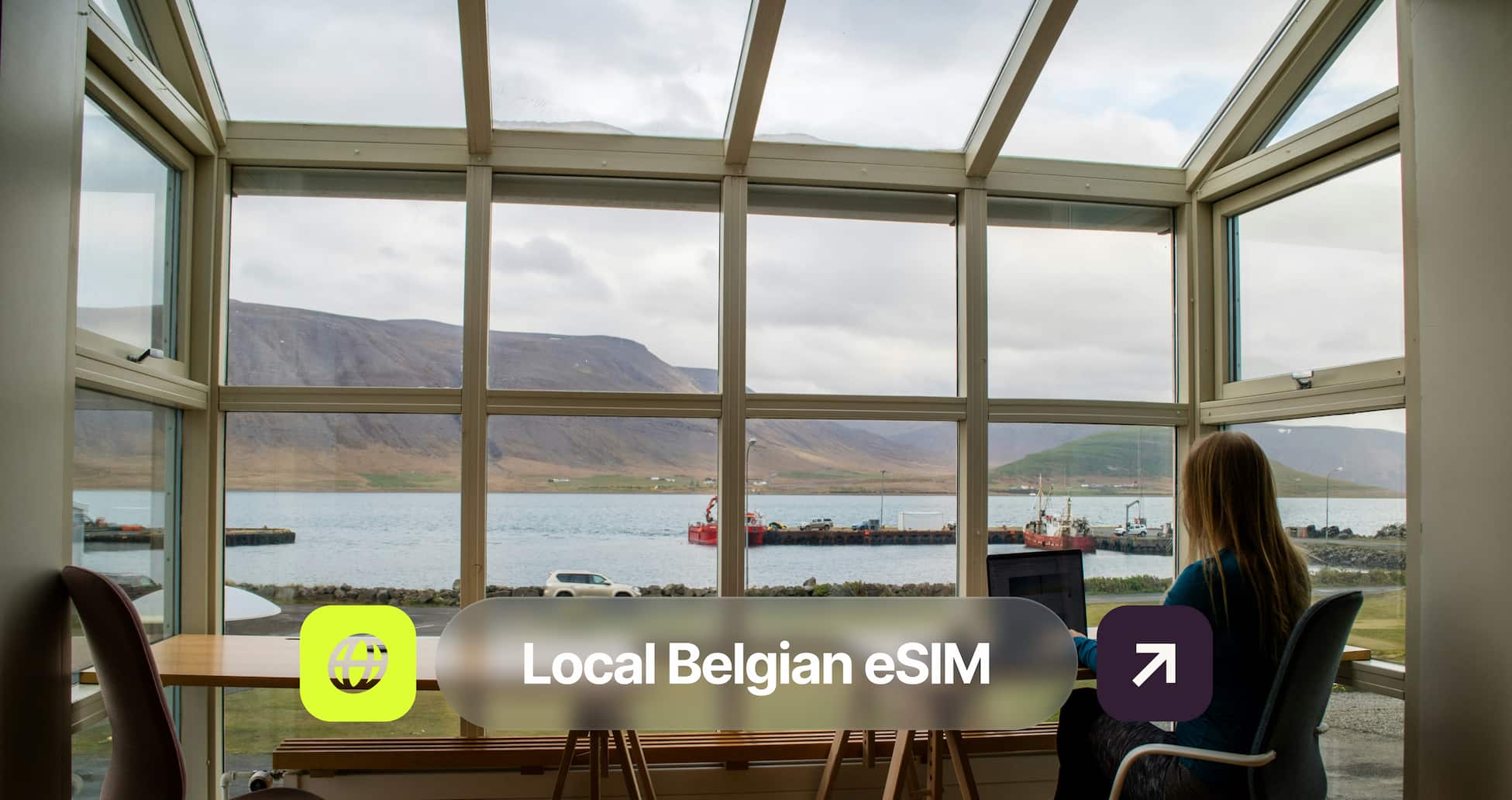 Person working on a laptop by a large window overlooking a scenic lake and mountains, with text overlay "Local Belgian eSIM."