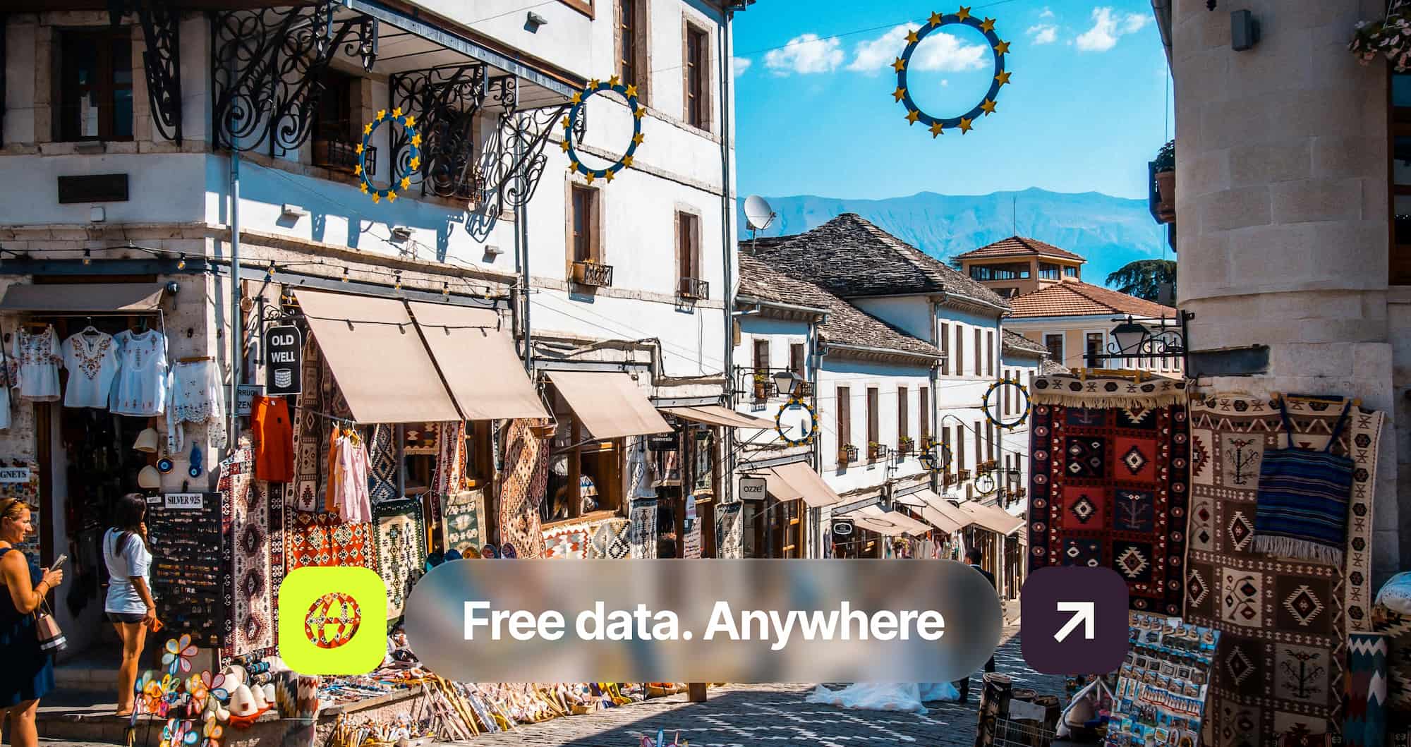 A street lined with traditional shops displaying colorful textiles and garments, under a clear blue sky with mountains in the background.
