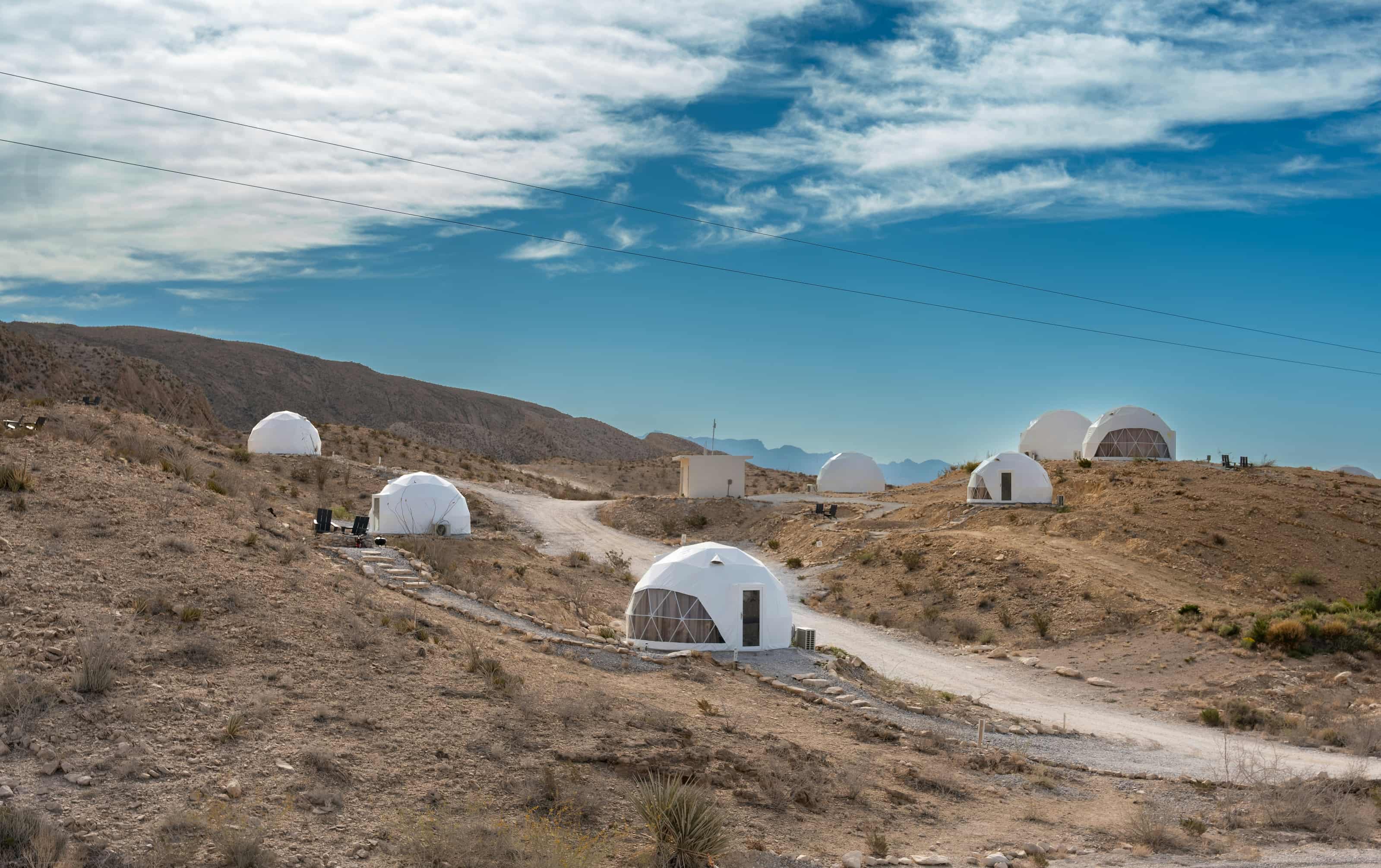 A desert landscape featuring several small, white dome structures scattered across sandy hills under a blue sky with wispy clouds.